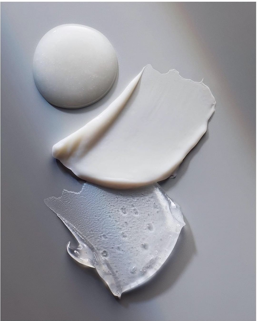 Close-up studio shot of a collection of skincare product textures, including a thick white cream, lotion smear, and clear gel, on gray.