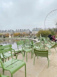 Rows of green metal park chairs surrounding a reflecting pool with a Ferris wheel and classical European buildings in the distance.
