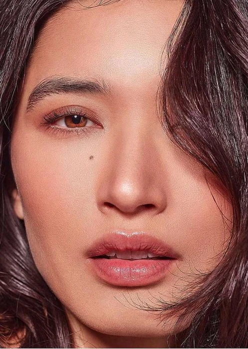 Close-up portrait of a woman with brown eyes, dark hair partially covering her face, and a beauty mark on her cheek.