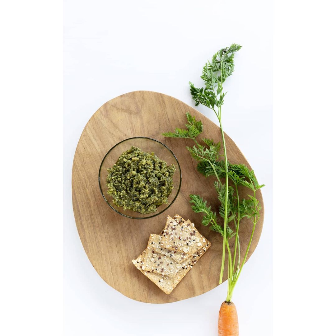 Overhead shot of a wooden board with a bowl of green spread, crackers, and a carrot with leaves on a white background.