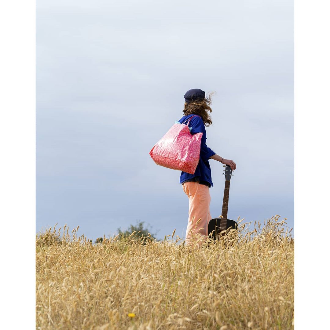 Young adult with guitar stands in a wheat field on a sunny day, wearing a blue shirt and peach-colored pants with a floral tote bag.