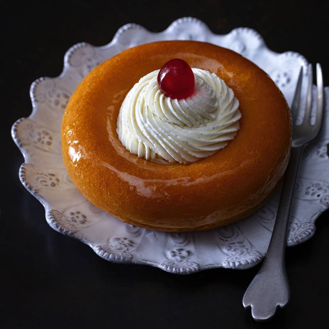 Close-up shot of a rum baba dessert on a decorative white plate with a fork, showcasing its glazed surface, cream topping, and cherry.