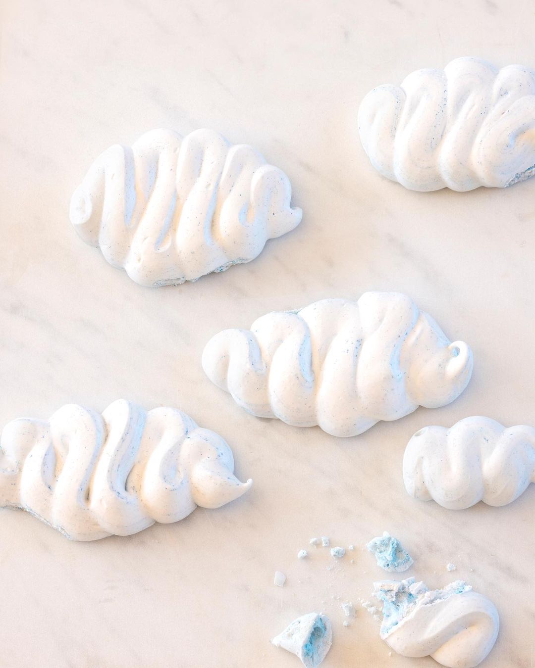 Five cloud-shaped meringues with white and blue coloring are scattered on a marble surface with one meringue partially broken.