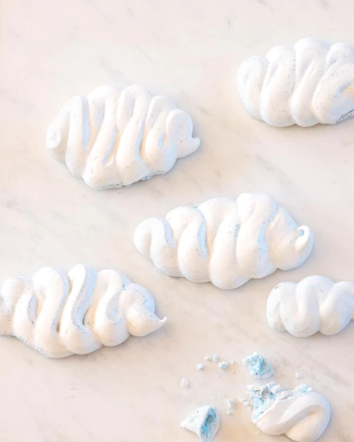 Five cloud-shaped meringues with white and blue coloring are scattered on a marble surface with one meringue partially broken.