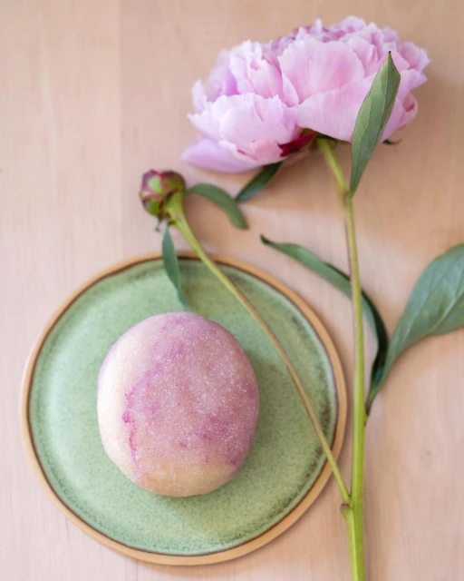 Overhead shot of a light purple mochi on a light green plate next to a pale pink peony bloom and bud against a pale wood background.