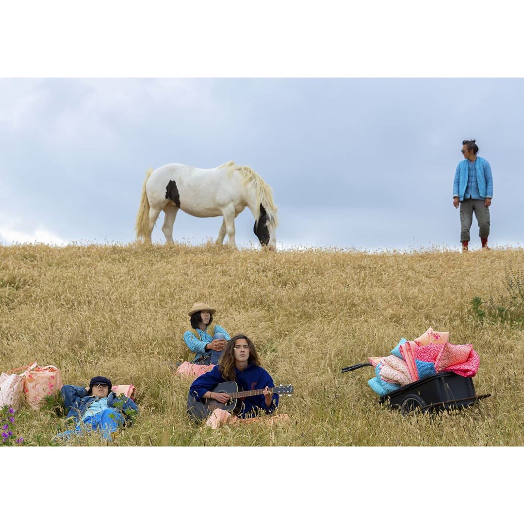Group of four people and a horse in a wheat field on a cloudy day. One man stands while the others relax. A cart of blankets sits nearby.