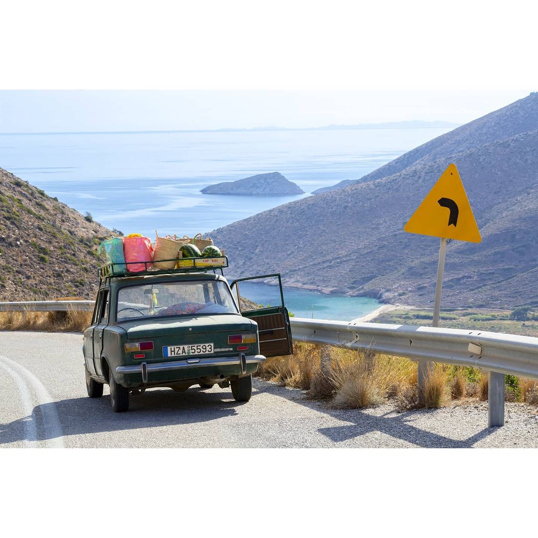 A vintage green car parked on a mountain road with ocean views, carrying luggage and watermelons on its roof rack, sunny day.