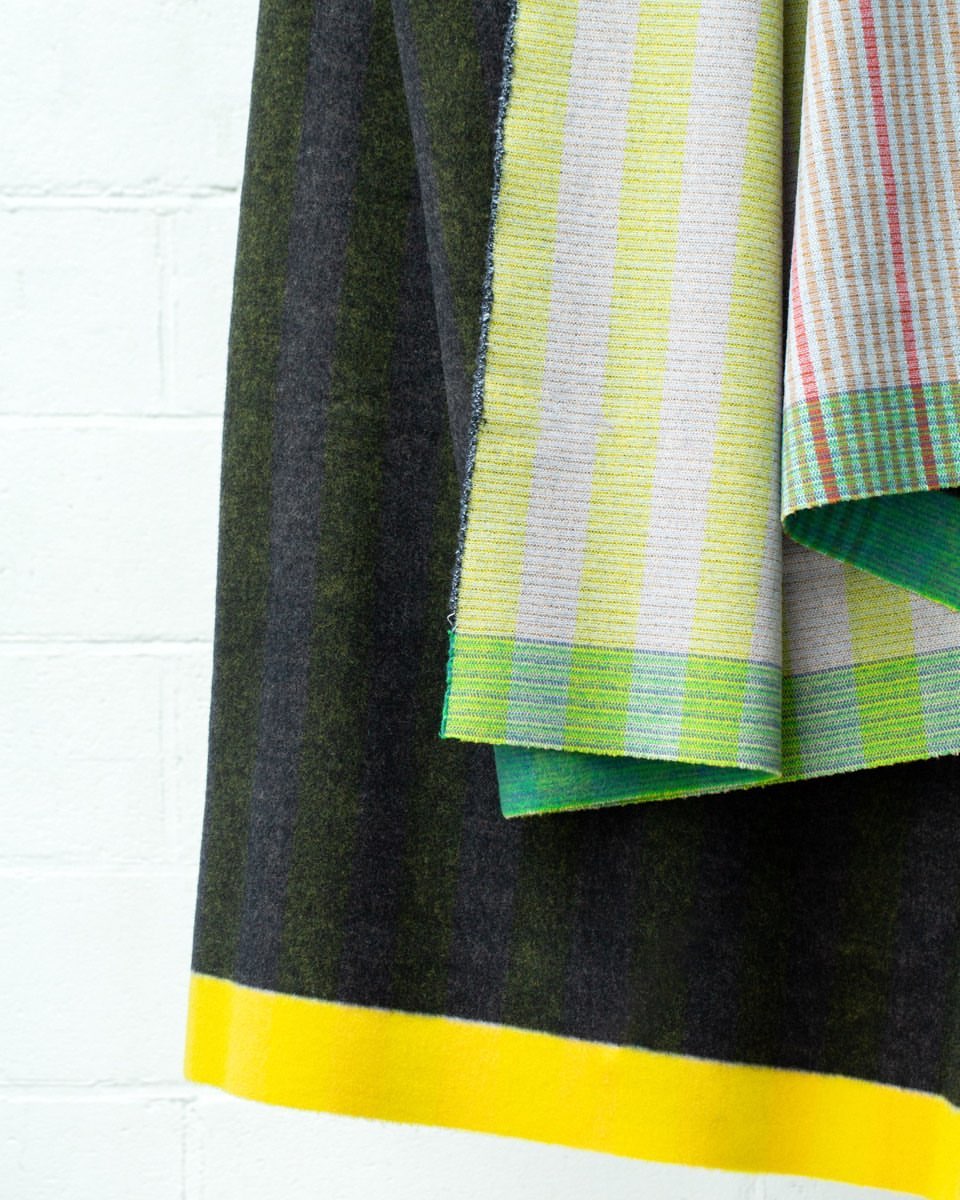 Close-up of folded fabrics with green, yellow, and gray striped patterns, alongside a white brick wall backdrop, creating a vibrant textile display.