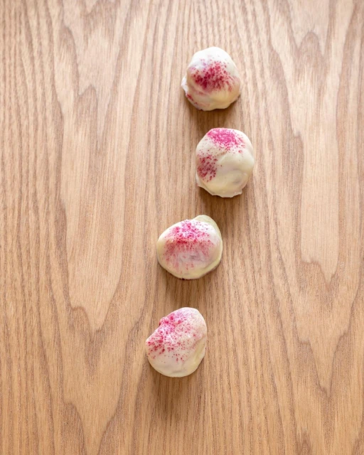 Overhead shot of four white chocolate truffles with pink sprinkles arranged on a light brown wooden surface.