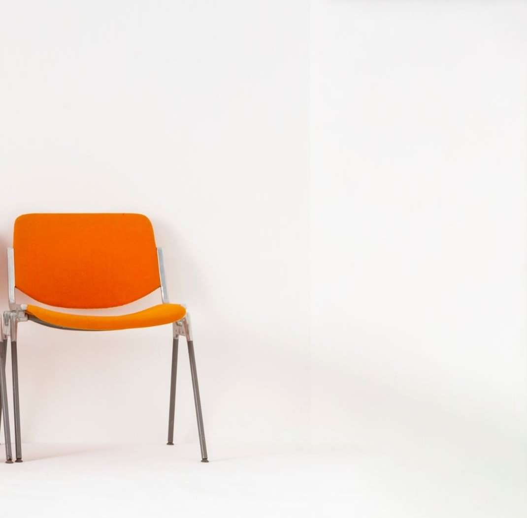 A single orange chair with a metal frame sits against a white wall, studio lit with ample negative space to the right.