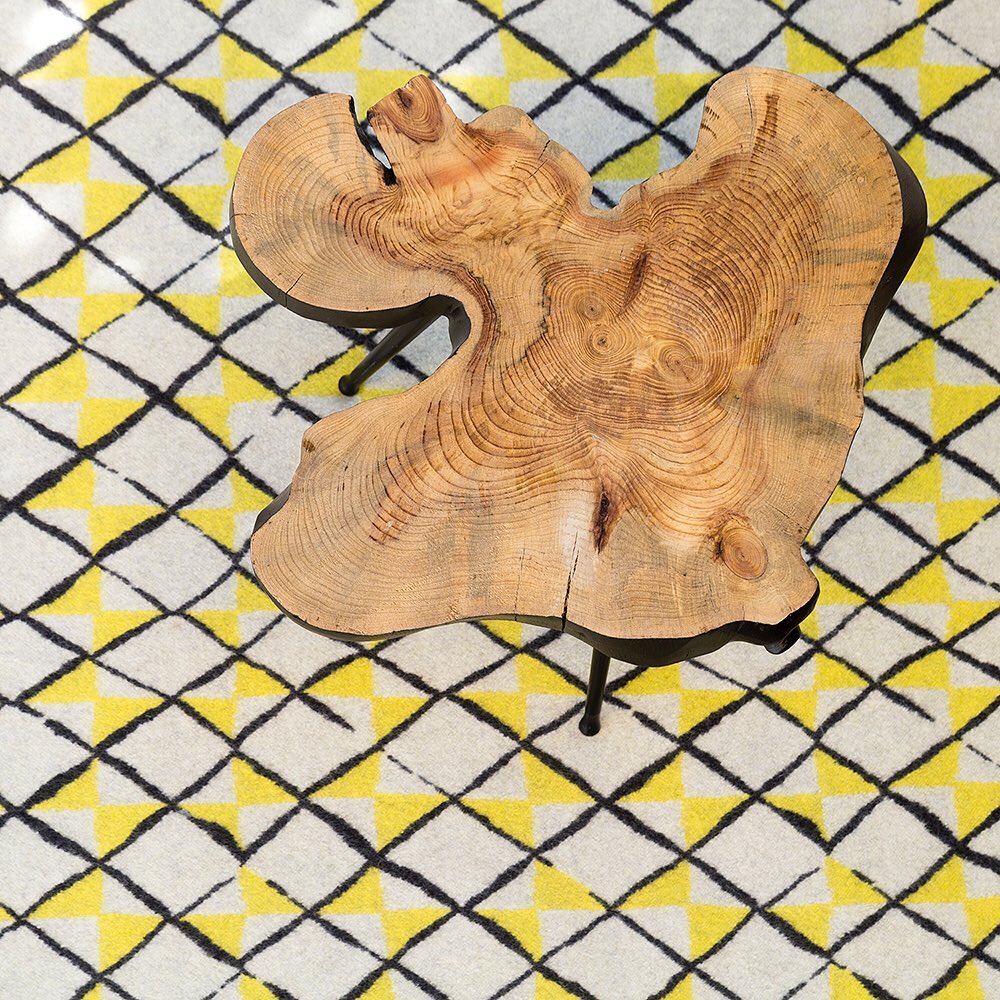 Elevated view of a round coffee table with a natural wood top and black metal legs on a patterned rug with yellow and black geometric shapes.