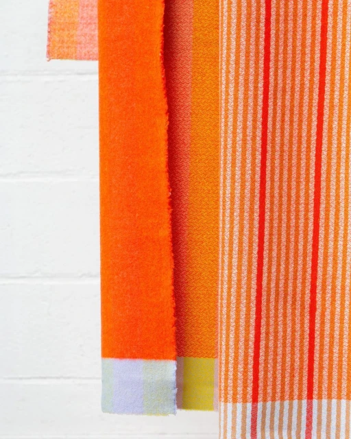 Close-up of folded orange and yellow fabric swatches with varying stripe patterns hanging against a white brick wall backdrop.