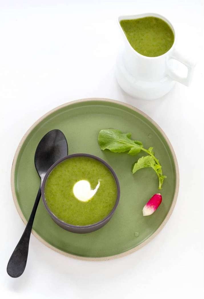 Overhead shot of vibrant green cream soup with a swirl of cream, served with a spoon, radish, and white ceramic pitcher.