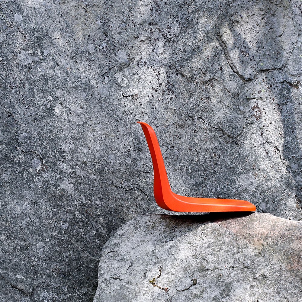 Orange chair-like structure positioned on a light gray stone, against a background of a textured gray stone wall in a natural outdoor setting.