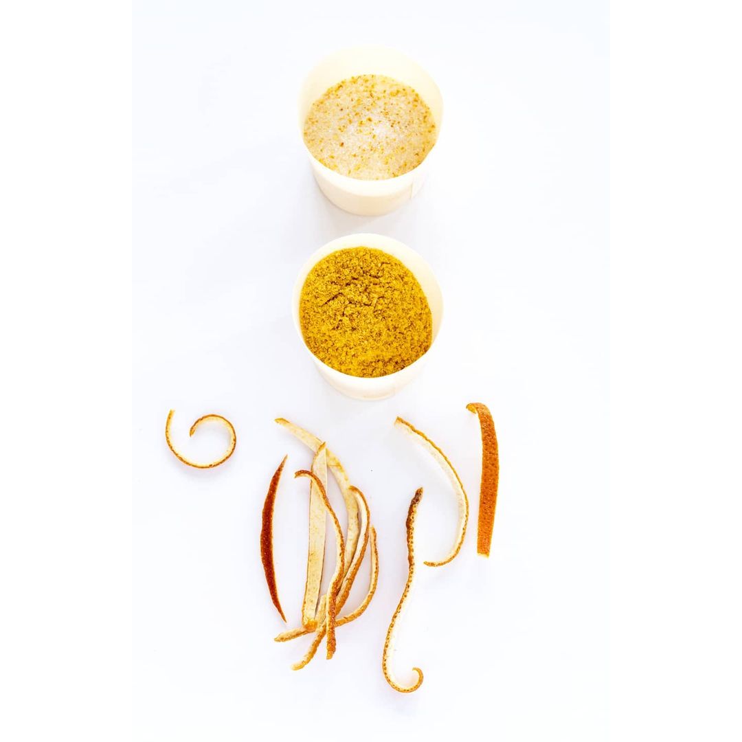 Elevated view of two white cups filled with light brown and golden yellow powder and several pieces of dried orange peel on white surface.