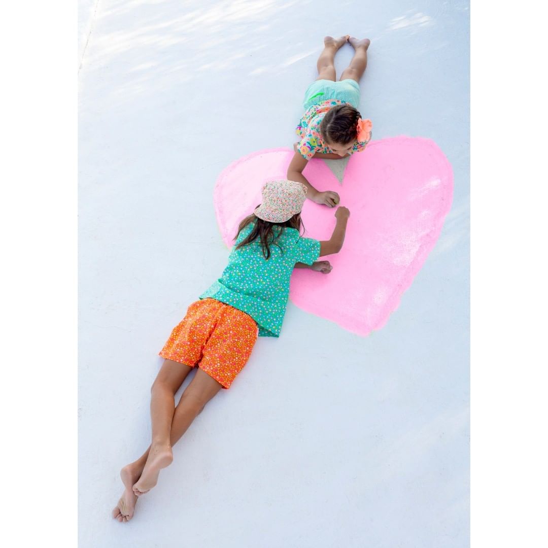 Two children lying on white surface next to a pink heart shape, wearing patterned clothing. Top down view, summer fashion.