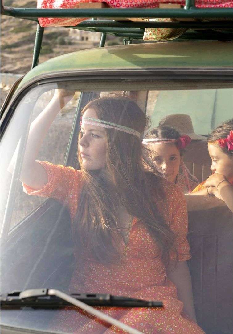 Woman driving a vintage green car with two children in the back seat on a sunny day. The car has a luggage rack on top.