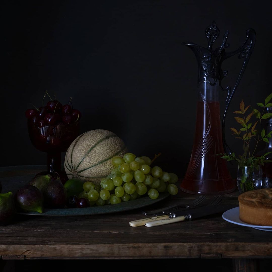 Still life photography of grapes, cherries, melon, cake, and antique silverware on a rustic wooden table against a dark background.