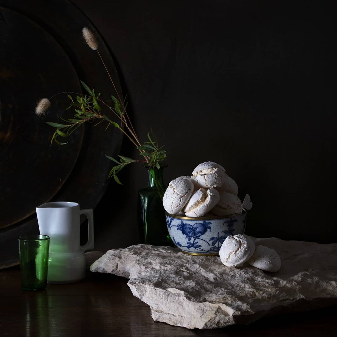 Still life featuring a bowl of white cookies, a green glass, a white mug, and a green-stemmed vase with plant sprigs on a stone slab.