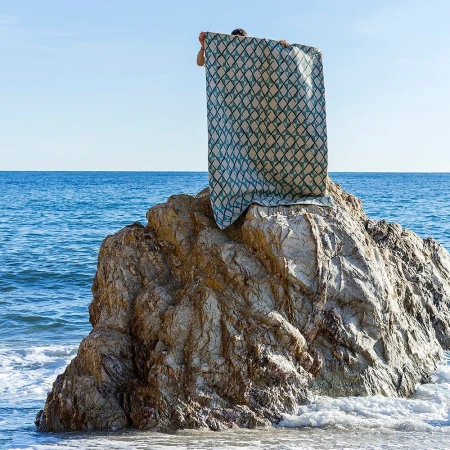Person holding a patterned textile banner atop a rocky outcrop by the ocean, photographed in daylight.