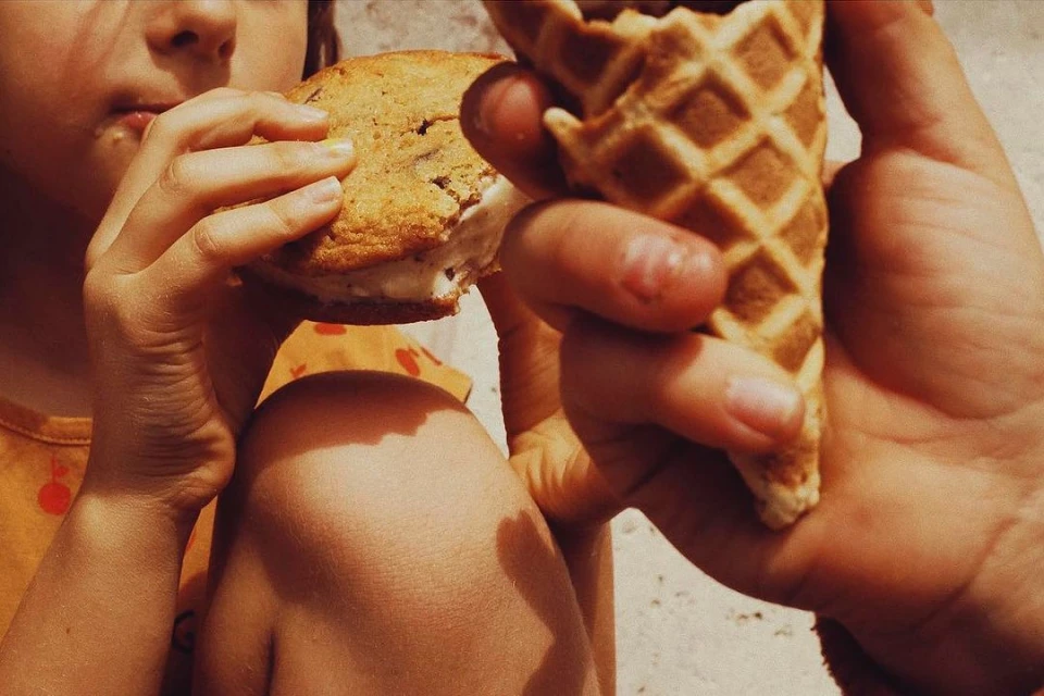 Child eating a cookie ice cream sandwich and another person holding a waffle ice cream cone. Close-up, food photography.