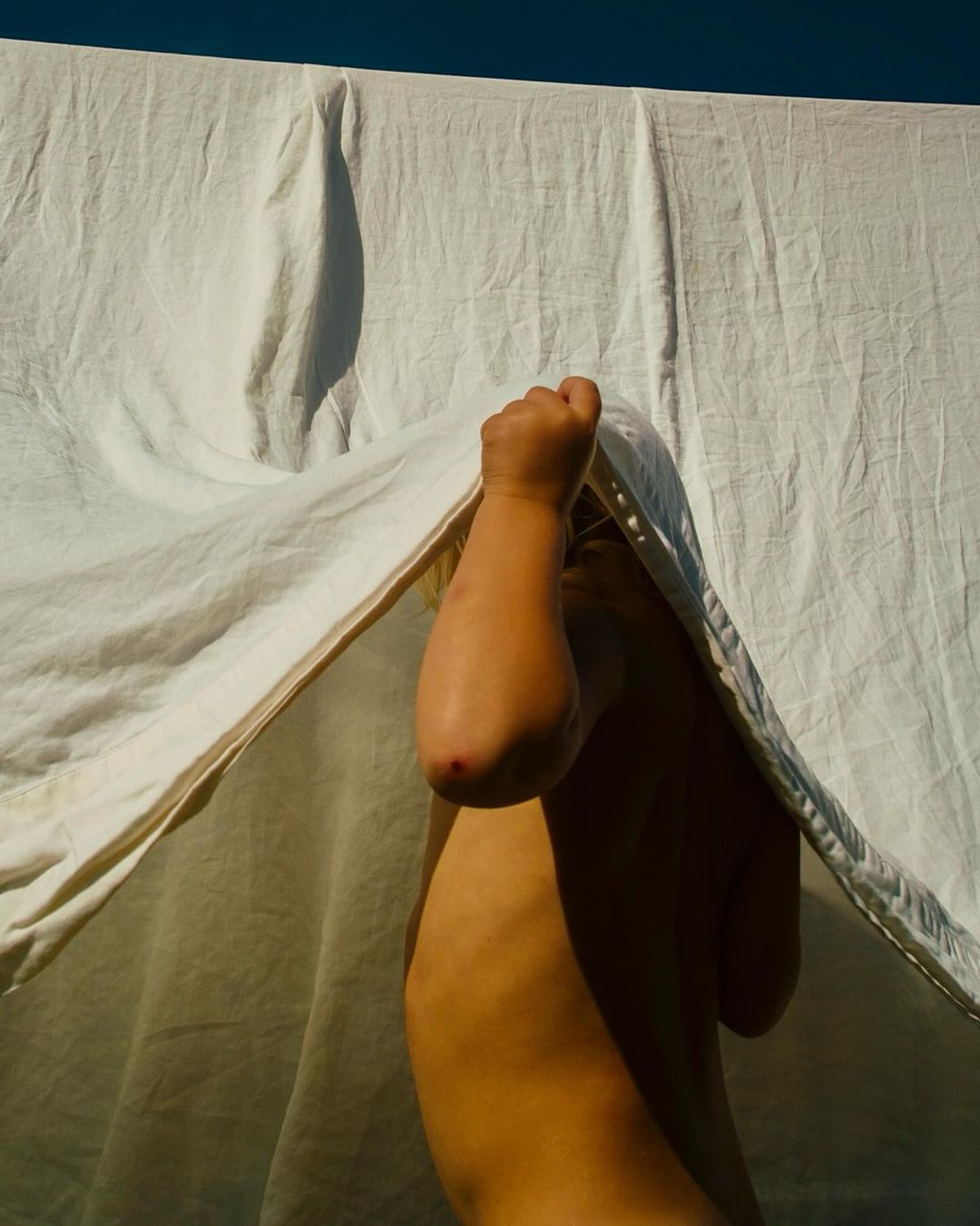 Child partially concealed behind a white cloth or sheet, bathed in natural sunlight, against a clear dark blue sky backdrop.