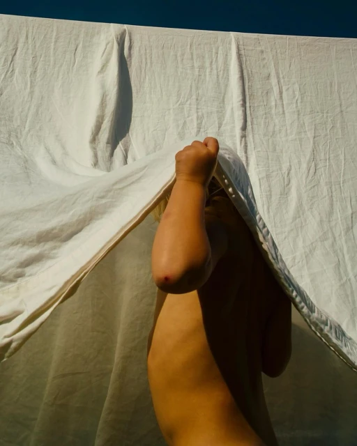Child partially concealed behind a white cloth or sheet, bathed in natural sunlight, against a clear dark blue sky backdrop.