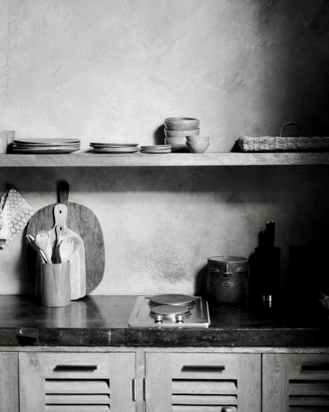 Rustic kitchen interior in monochrome featuring wooden shelves, tableware, cooking utensils, and textured wall, creating a natural ambiance.