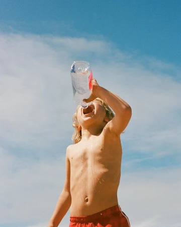 Child with blonde hair drinking water from a clear plastic bottle against a blue sky, captured from a low angle by photographer Claire Guarry.