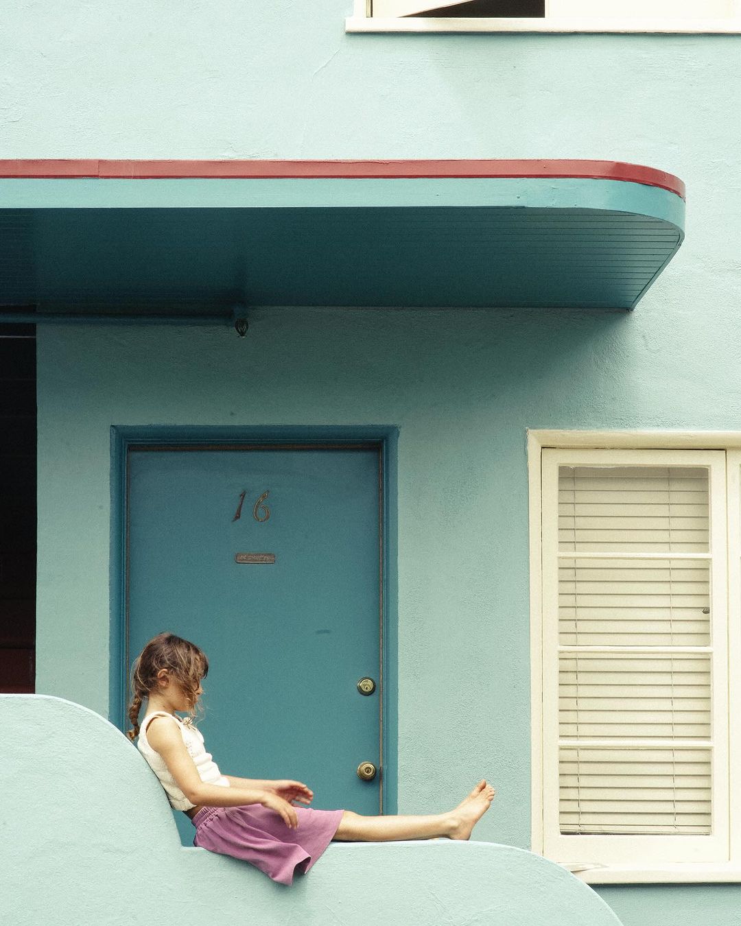 Young girl sits on the steps of a pale-blue building with a matching door and a white-framed window during daytime hours.