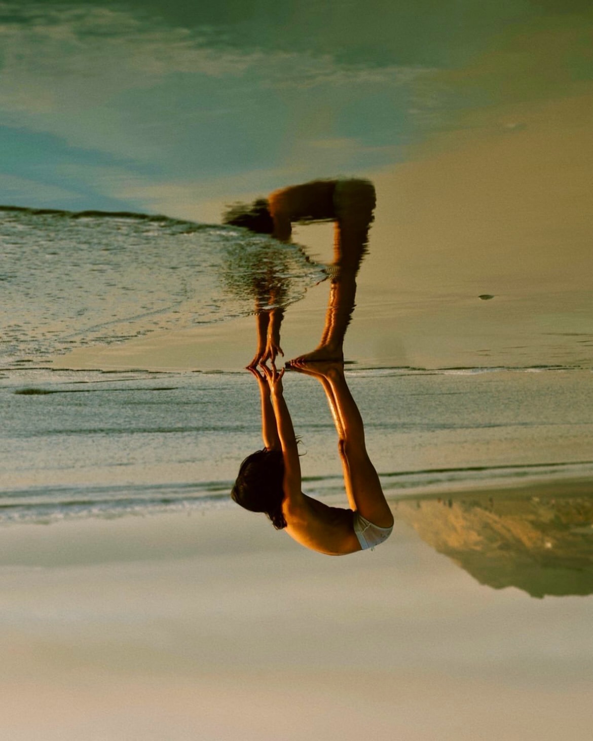 Woman bending over at a beach with reflection creating optical illusion effect. Sand, water, and blue sky blend at sunset.