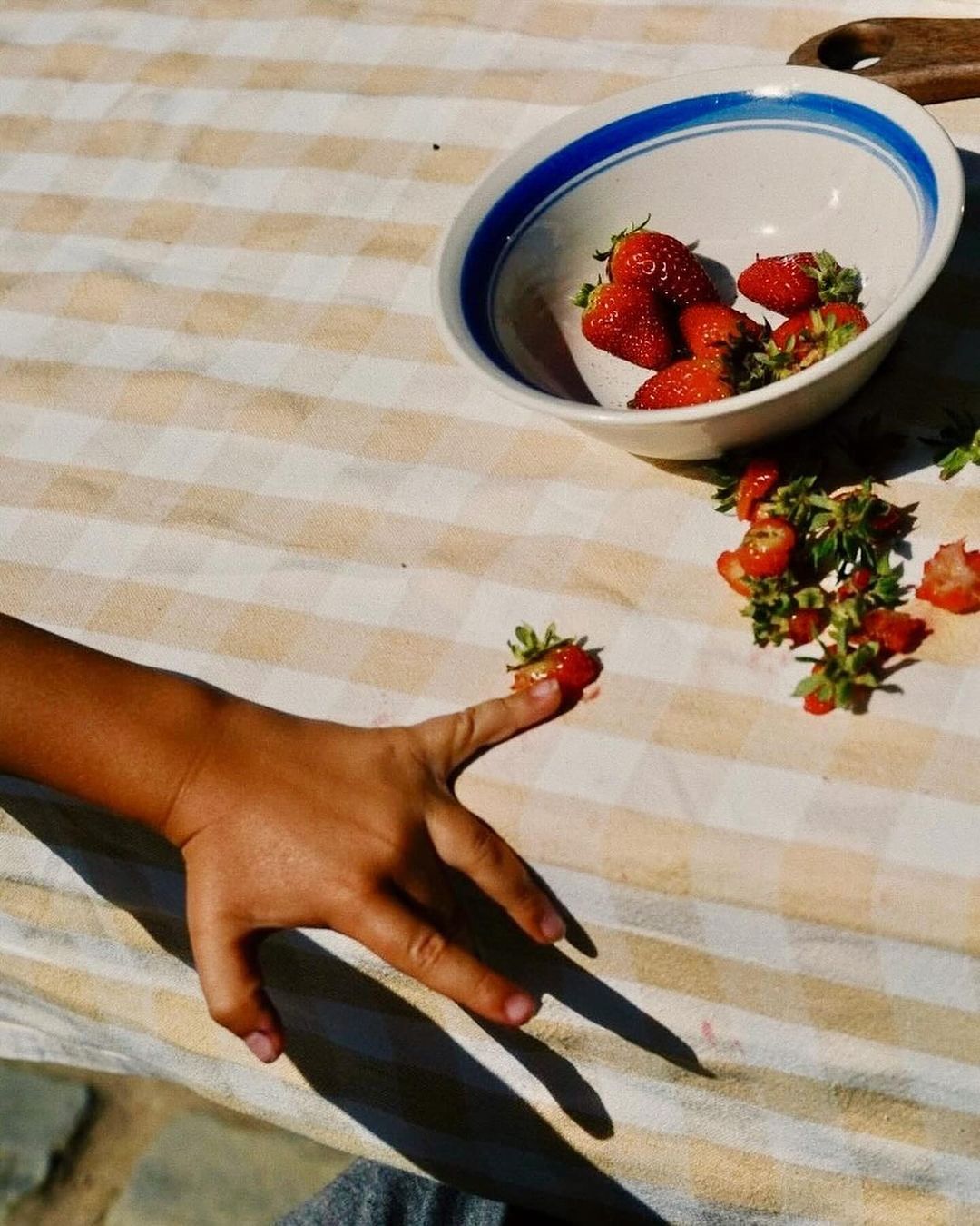 A child's hand holds a strawberry on a finger, next to a bowl of strawberries on a striped tablecloth, under natural light.