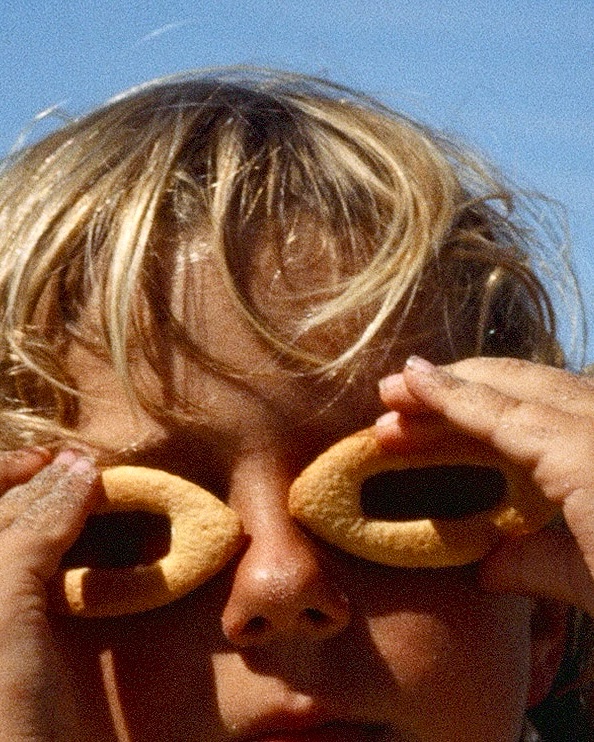 Young child playfully poses with two donut-shaped cookies held up to their eyes, set against a clear blue outdoor sky backdrop.