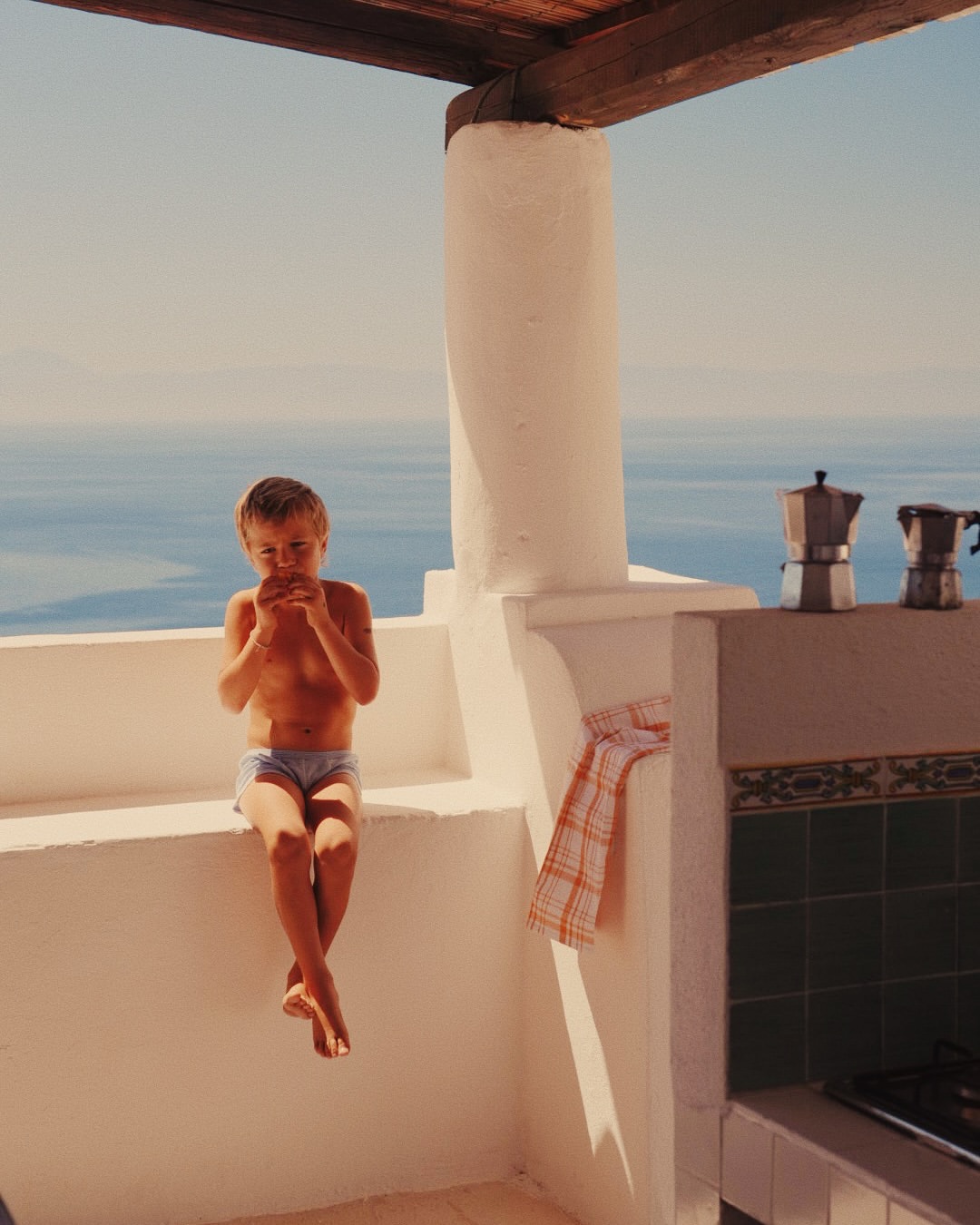 Young boy eating on a sunlit terrace with a sea view in the background, capturing a moment of leisure and scenic beauty.