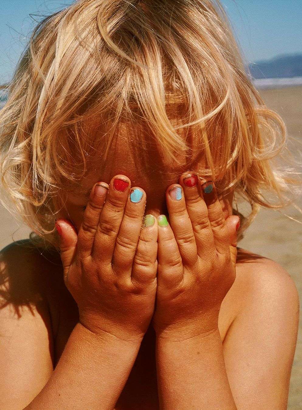 Child covers face with hands painted with different color nail polish on a beach with water and sand in the background during daytime.