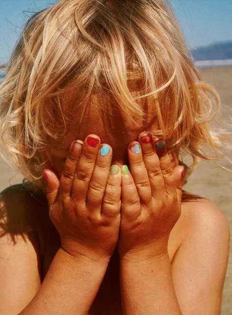 Child covers face with hands painted with different color nail polish on a beach with water and sand in the background during daytime.