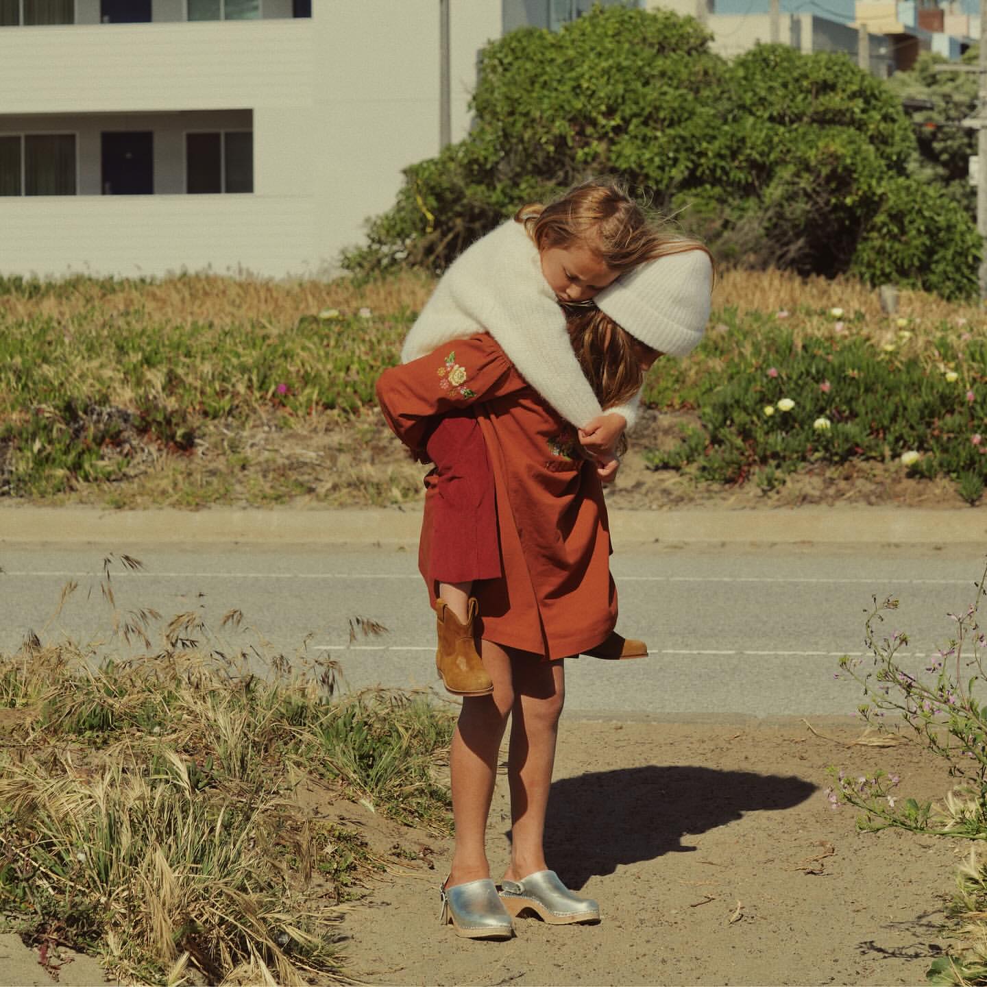 Young girl piggybacking a smaller child on a road near a grassy area with residential buildings, both wearing neutral tones.