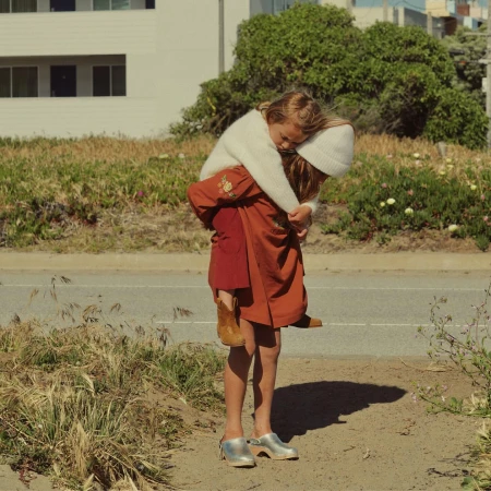 Young girl piggybacking a smaller child on a road near a grassy area with residential buildings, both wearing neutral tones.