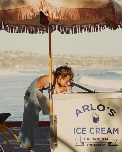 Child leaning over Arlo's Ice Cream cart near the coast on a sunny day, with a vintage umbrella and a bicycle attached.