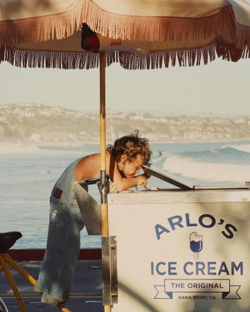 Child leaning over Arlo's Ice Cream cart near the coast on a sunny day, with a vintage umbrella and a bicycle attached.