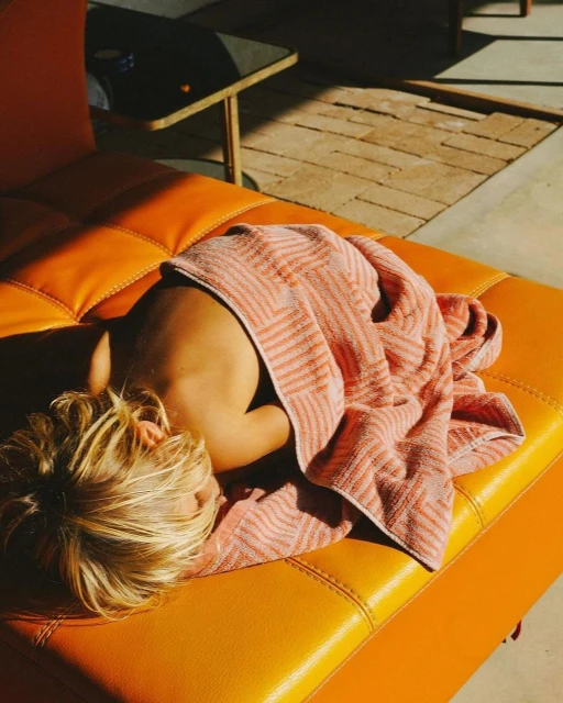 Child sleeps on an orange leather couch wrapped in a peach-colored striped towel on a sunny patio with visible brick flooring.