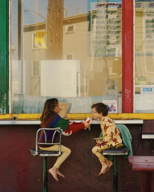 Two children sit at a colorful outdoor burger counter, eating and interacting in bright sunlight, with colorful signage visible.