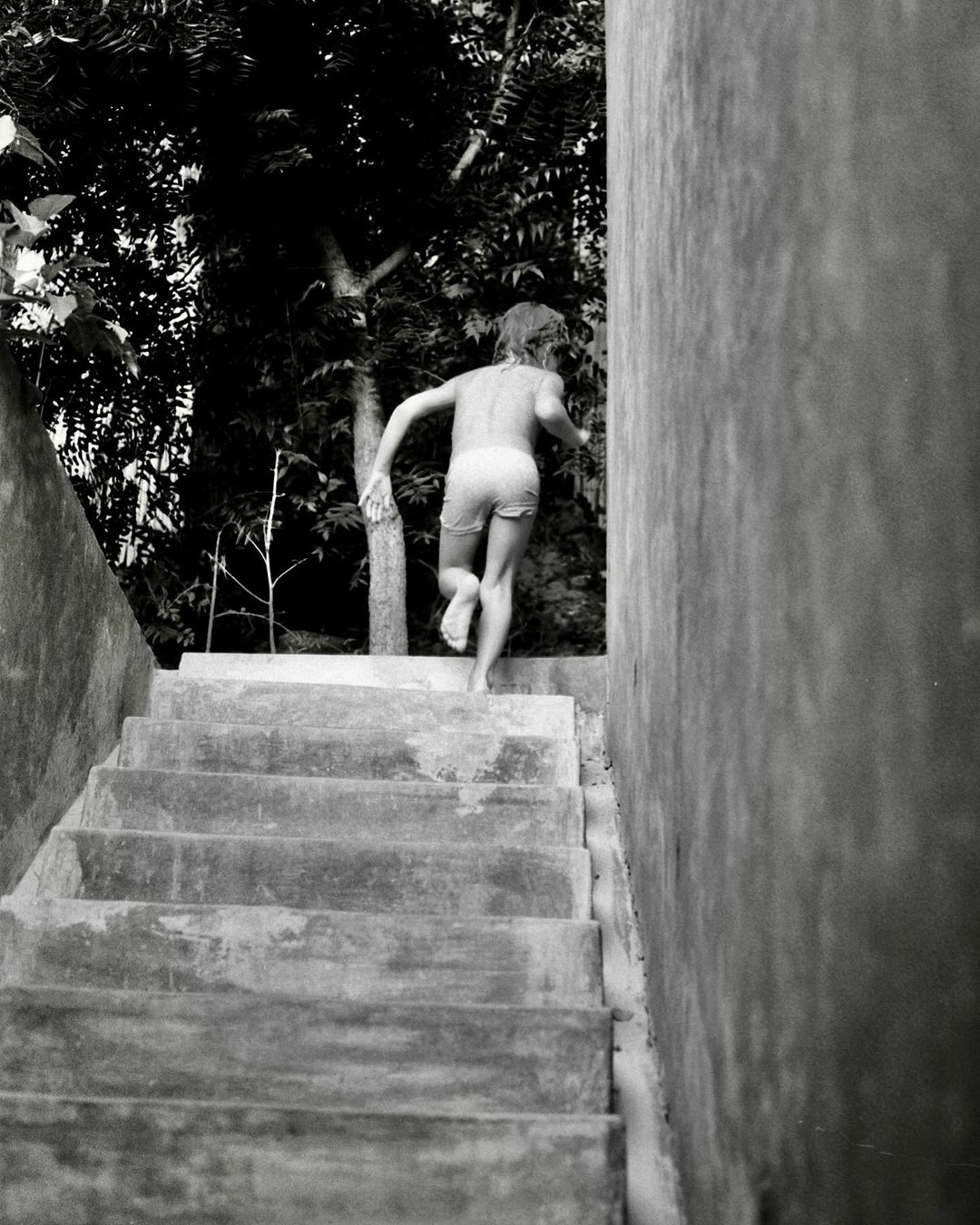 Black and white photo of a child climbing concrete stairs outdoors, with lush foliage and a tall concrete wall in the background.
