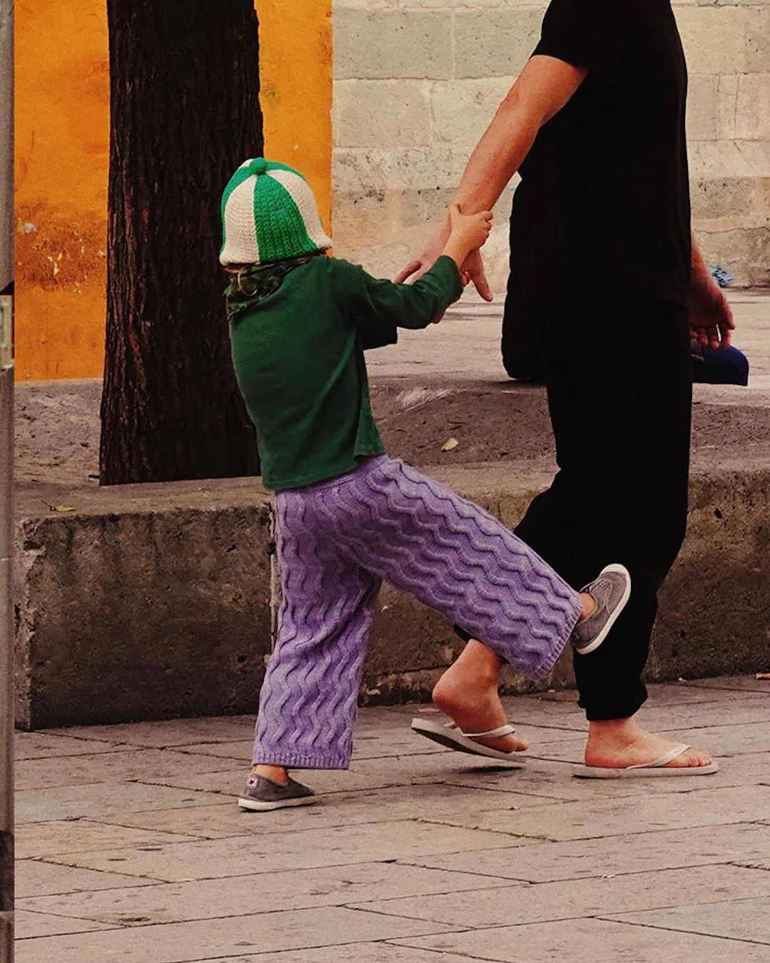 Child in purple pants and green sweater pulls away from an adult on a city sidewalk with orange wall and tree, holding hands.