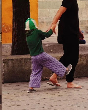 Child in purple pants and green sweater pulls away from an adult on a city sidewalk with orange wall and tree, holding hands.