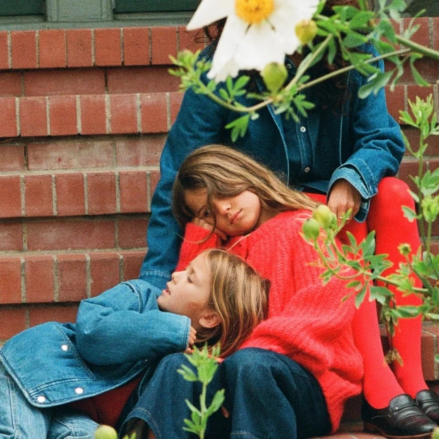 Three young girls wearing denim and red outfits sitting together on brick steps with flowers and greenery in an outdoor setting.