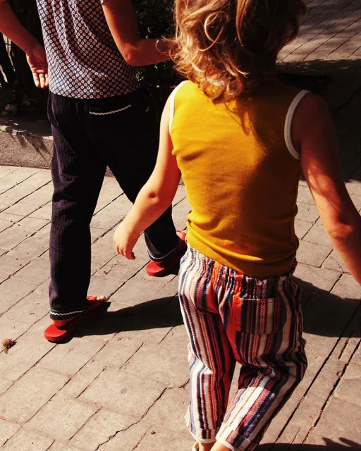 Young child in yellow tank top and striped pants walking with adult on brick surface with shadows, sunny, outdoors, daytime.
