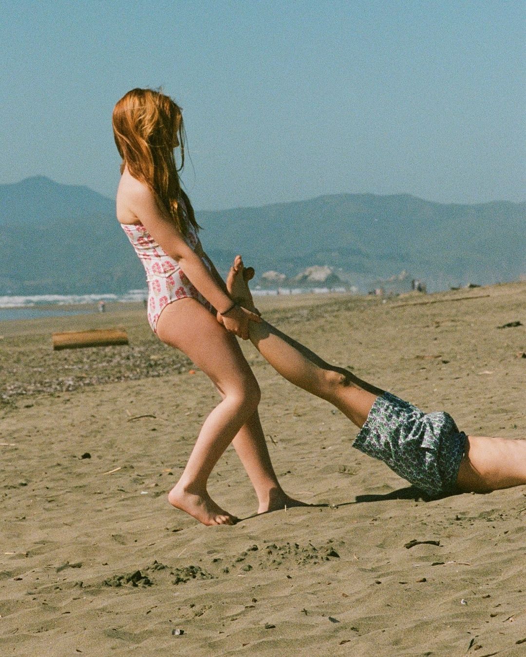 Girl in swimsuit pulls on legs of another person lying on sand at sunny beach, ocean and distant mountains in the background.