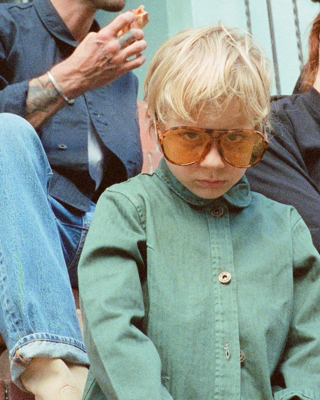 Child in vintage sunglasses, green jacket, and blonde hair. Nearby adults eating. Casual family moment with a retro aesthetic.