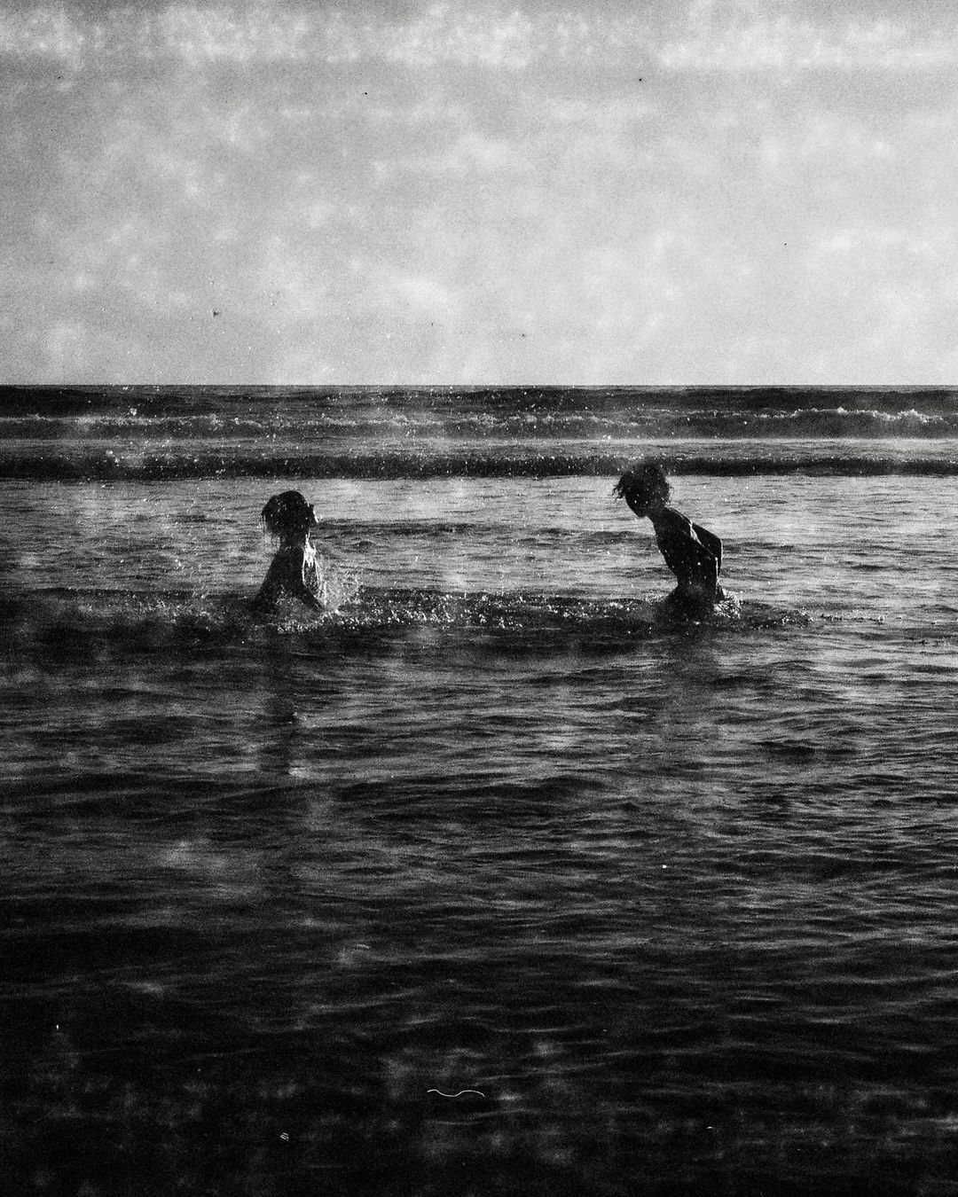 Two children wade in the ocean surf, their silhouettes against the horizon, rendered in grayscale with visible film grain and texture.